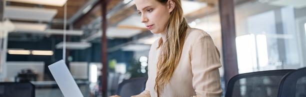 Mulher de negócios concentrada trabalhando em um laptop em um moderno espaço de escritório de conceito aberto, representando a produtividade e o trabalho digital.