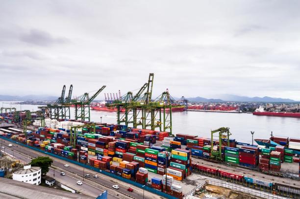 Vista aérea de porto marítimo com contêineres coloridos, guindastes e navios à beira-mar