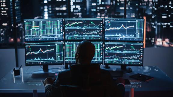 Man sitting facing window looking at 6 screens of financial data