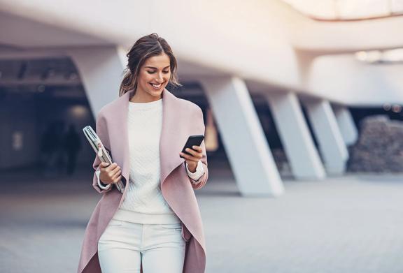 Stylish young professional woman walking outdoors, holding a newspaper and smiling while checking her smartphone.