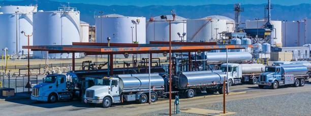 Fuel tanker trucks lined up at a refinery or petroleum distribution terminal with large storage tanks in the background.