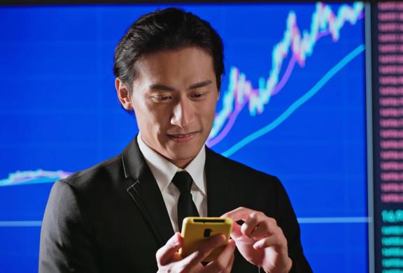 Business professional using a smartphone in front of a large screen displaying financial charts and stock market data.