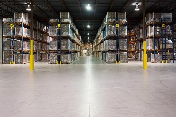 Interior view of a spacious industrial warehouse featuring tall pallet racks filled with stacked inventory boxes, well-lit aisles, and a clean concrete floor.