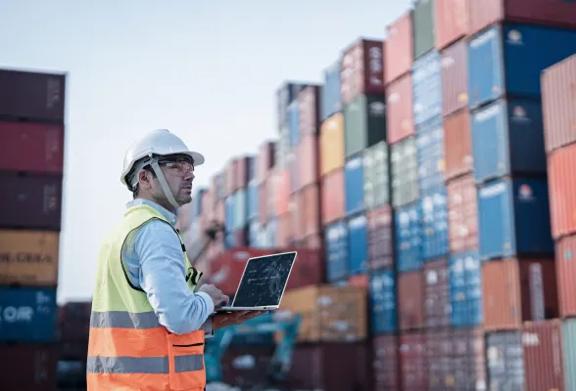 Trabalhador de logística usando laptop para inspecionar contêineres empilhados no terminal portuário.