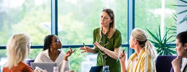 Four women in an office meeting, with one standing and gesturing while the others listen, surrounded by large windows and greenery.