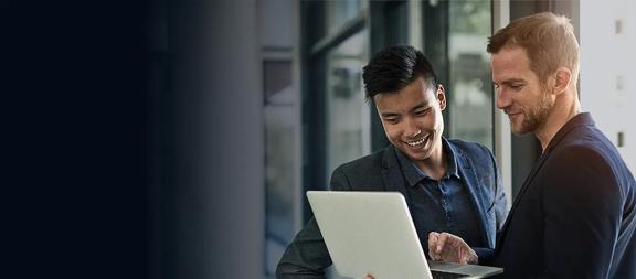 Dois profissionais em frente a um notebook prateado, discutindo informações em ambiente corporativo com parede de vidro.