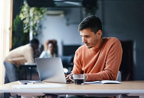 Man in orange sweater working on laptop at desk with notebook and glass of coffee in modern office
