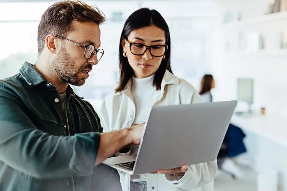 Duas pessoas analisando informações em notebook prateado, em ambiente claro com prateleiras ao fundo.