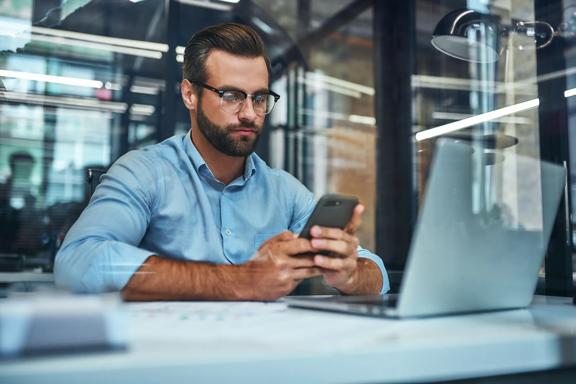 Focused businessman using smartphone while working at modern office desk with laptop