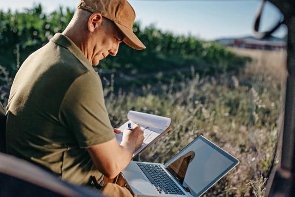 Agricultor sentado ao ar livre, escrevendo em caderno e utilizando laptop para registrar dados da lavoura.