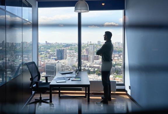 Businessman standing in modern office with arms crossed, overlooking cityscape through large window during daytime.