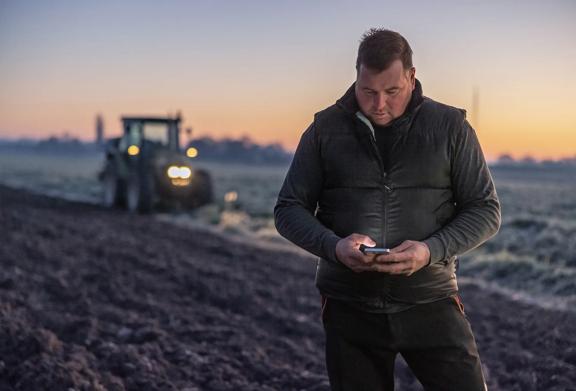 Farmer standing in field looking at his mobile phone with a tractor in the background
