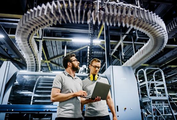 Two workers using a laptop in a manufacturing facility with overhead machinery.