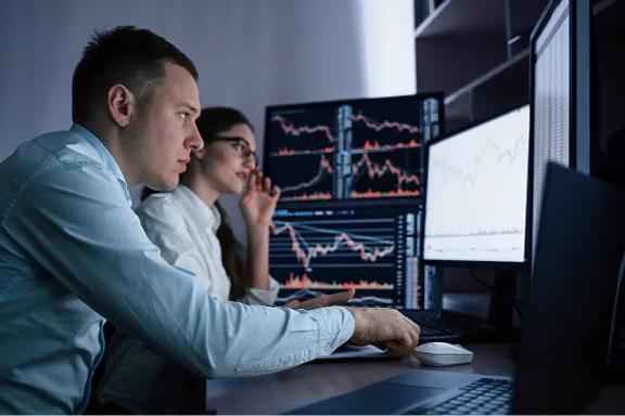 Traders analyzing financial market charts on multiple monitors in an office.