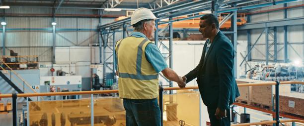 A factory manager in a safety vest and hard hat shaking hands with a business professional in a suit inside a large industrial manufacturing facility, symbolizing collaboration and partnership.