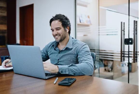 Homem sorrindo enquanto trabalha em seu laptop em um escritório moderno.