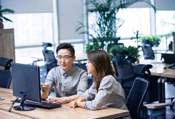 Man pointing at computer monitor while discussing work with woman in modern office with plants 