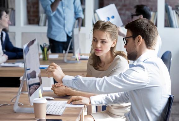 Man pointing to computer monitor while discussing work with a woman at an office desk, coffee cup on table