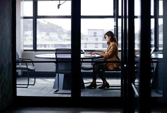 A businesswoman working alone on a laptop in a modern glass-walled conference room with cityscape views in the background.