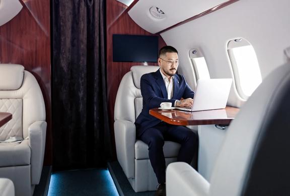 Businessman working on laptop in luxury private jet cabin, seated at wood-finished table with coffee cup.