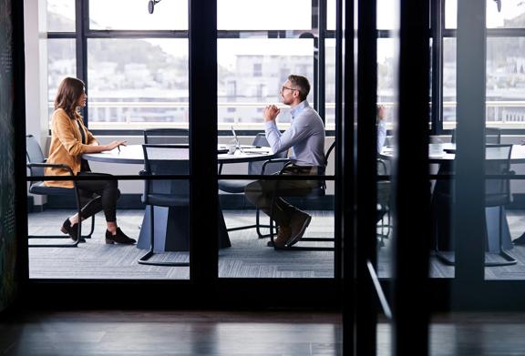 Businesswoman and businessman in discussion during formal meeting inside modern glass-walled conference room.