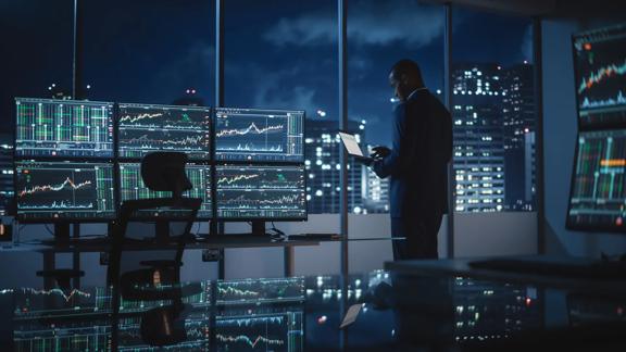 Man standing by window holding laptop next to multiple screens