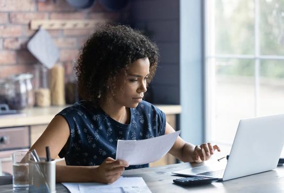 A woman working at a kitchen table with financial documents, a calculator, and a laptop, concentrating on budgeting or accounting tasks in a home office setting.
