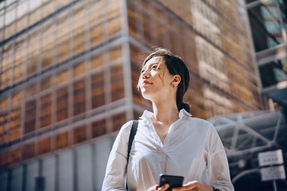 Person standing outdoors in front of a tall glass building, holding a smartphone and looking upward.