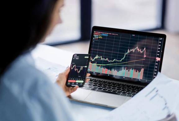 Trader analisando tendências do mercado de ações em um smartphone e laptop com gráficos financeiros