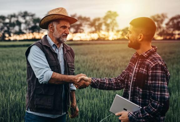 A senior farmer in a straw hat shaking hands with a younger professional holding a tablet, standing in a green crop field during sunset, symbolizing agricultural collaboration and trust.