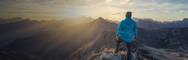 Homem em pé no cume de uma montanha ao nascer do sol, com vista para uma dramática paisagem alpina e picos iluminados pelo sol.