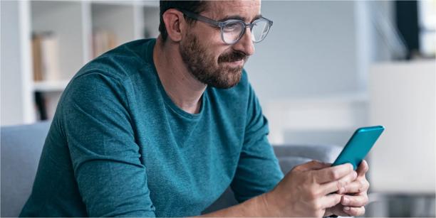 Person sitting indoors looking at a smartphone, with a laptop and shelves visible in the background.