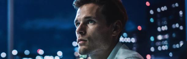 Thoughtful young professional looking out in a high-rise office at night with city lights in the background