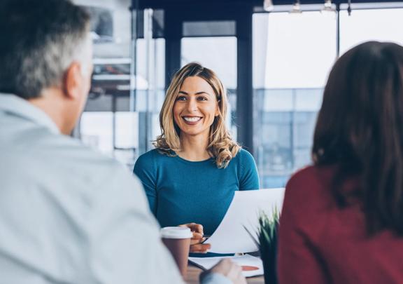 Mulher sorridente em uma reunião de negócios, segurando papéis e interagindo com colegas
