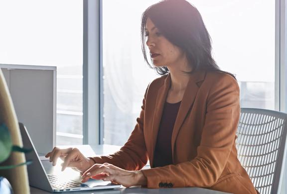 Focused businesswoman in a brown blazer working on a laptop at a sunlit office desk.