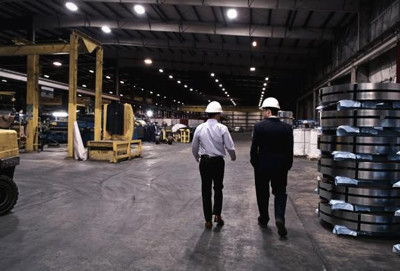 Two men wearing safety helmets walking through large industrial warehouse with machinery and stacked metal coils