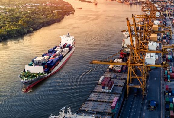 Navio de carga transportando contêineres por um porto com guindastes descarregando mercadorias.