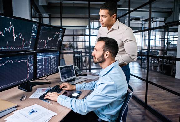 Two men analyzing financial charts on multiple monitors at a trading desk in modern office