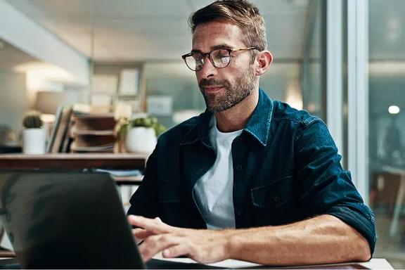 Profissional usando camisa casual azul sobre camiseta branca, trabalhando em notebook preto em escritório com prateleiras e plantas ao fundo
