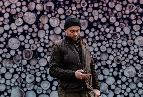 Man in outdoor workwear using a smartphone while standing in front of a large stacked pile of cut logs