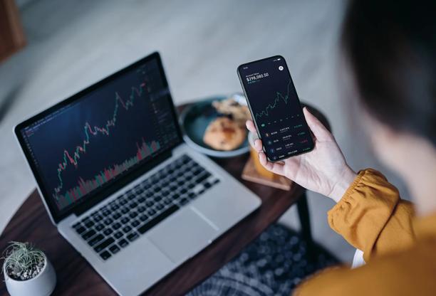 スマートフォンで金融チャートを表示し、木製テーブルの上には同様の株式市場データを表示するノートパソコンがある。