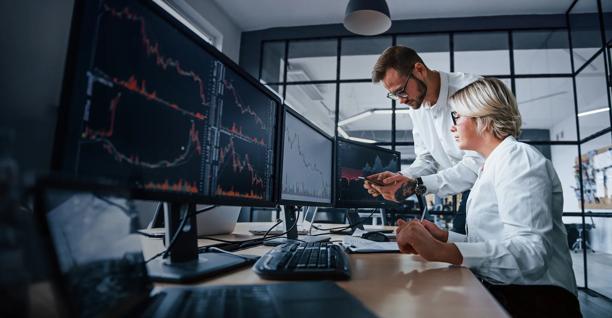 Um homem de pé e uma mulher sentada olhando dados do mercado financeiro exibidos em várias telas