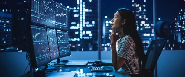 Focused woman analyzing stock charts on multiple monitors in a high-rise office at night