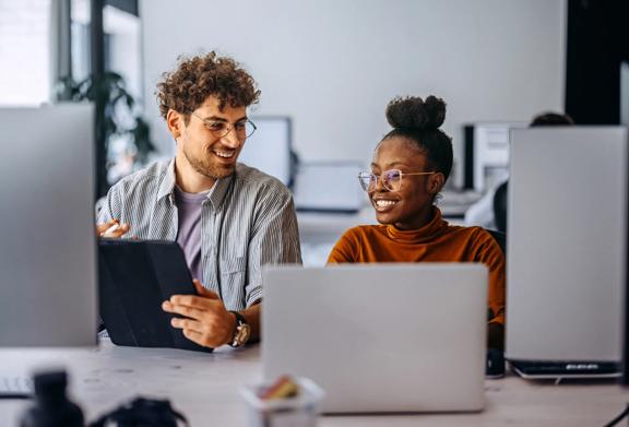 Man and woman collaborating at desk with laptops and digital tablet in modern office