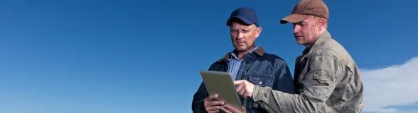 Two farmers outdoors under a clear blue sky using a tablet to review farm data and agricultural technology