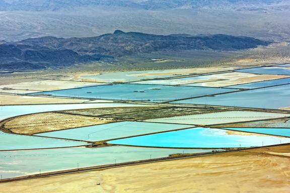 Aerial view of lithium evaporation ponds in a desert landscape, showing turquoise and blue pools divided by thin barriers with mountains in the background.