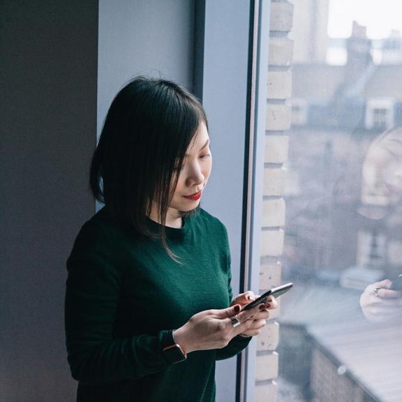Person standing by a window using a smartphone, with city buildings visible outside.