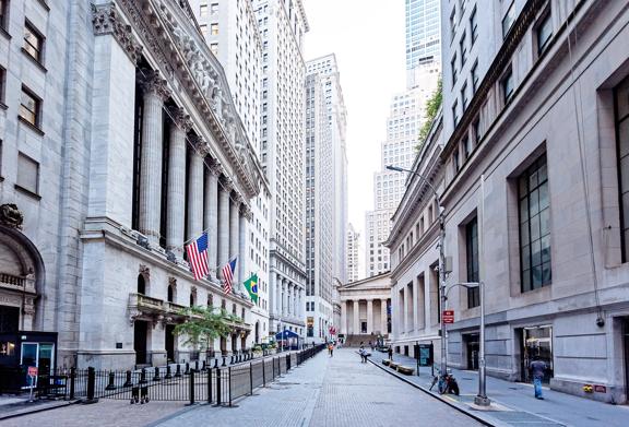 View of Wall Street in New York City with the New York Stock Exchange building and American flags on a bright day.