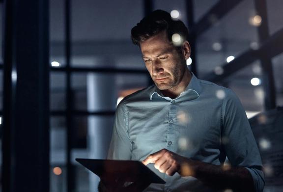 Business professional working on a digital tablet at night in a modern office setting, illuminated by city lights.