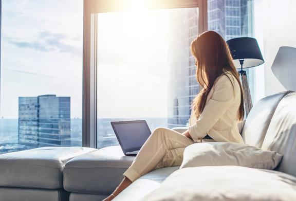 Businesswoman in white suit working remotely on a laptop in a modern high-rise apartment with city skyline views at sunrise.